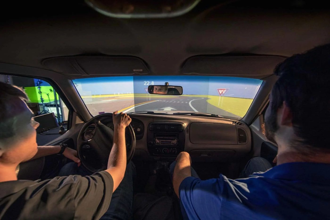Two students in a driving simulator cab driving a simulated roadway projected ahead
