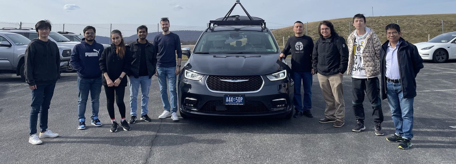 A group of lab members and visitors standing on either side of the lab's automated vehicle in a parking lot